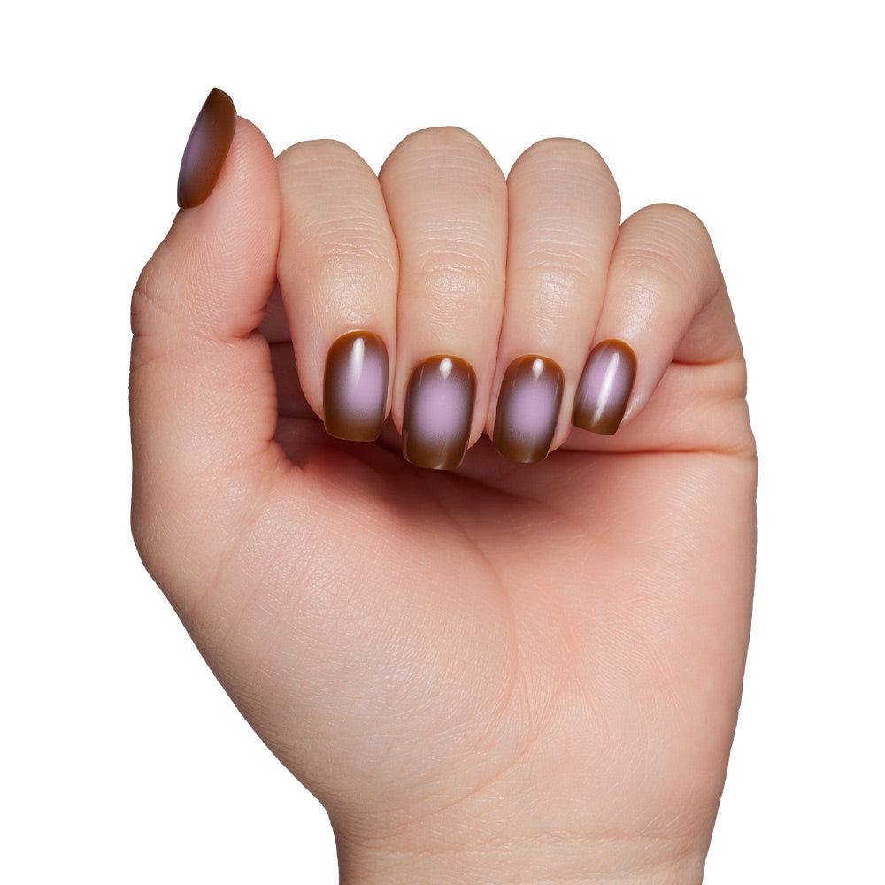 A hand with slightly curled fingers displays short squoval nails painted in a taupe brown and purple metallic gradient from the Eclipse set by glamnetic, shown against a white background. - Product image