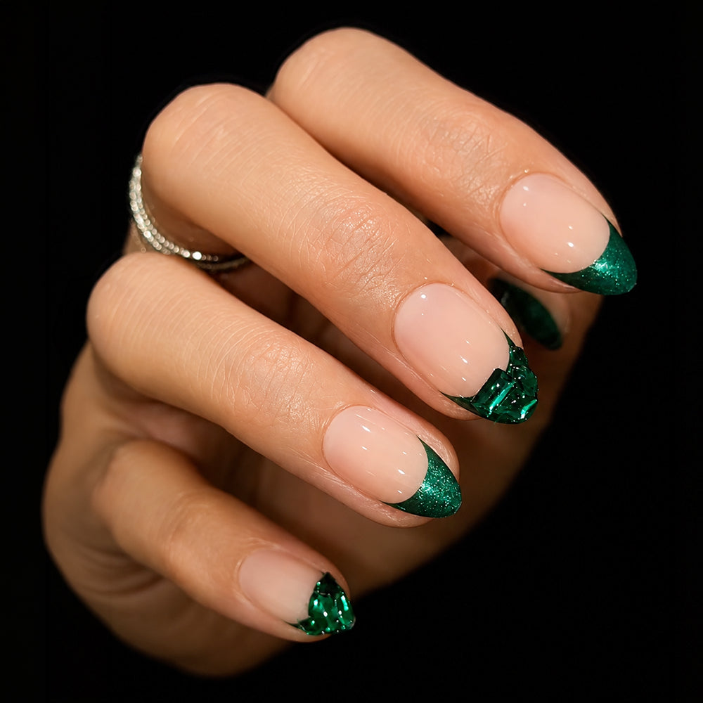 A hand with almond-shaped nails is styled with glamnetic's Emerald press-on nails, featuring a nude base and glittery emerald green French tips. Some nails are embellished with green foil accents, giving a gemstone-inspired look. The person accessorizes with a silver ring.