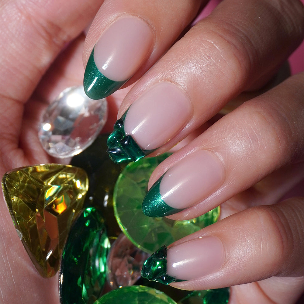 A hand with long, almond-shaped glamnetic Emerald Press-On Nails, styled with a green French tip manicure, holds several large, faceted gemstones in green and yellow tones. - Product image