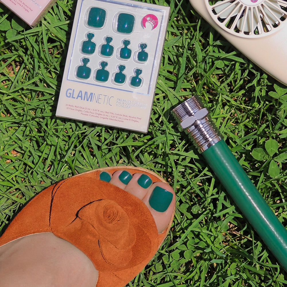 A foot in a tan sandal with a floral detail wears glamnetic Emerald Green Toenail press-ons and stands on grass. Nearby are glossy press-on toenails, a garden hose, and a portable fan.