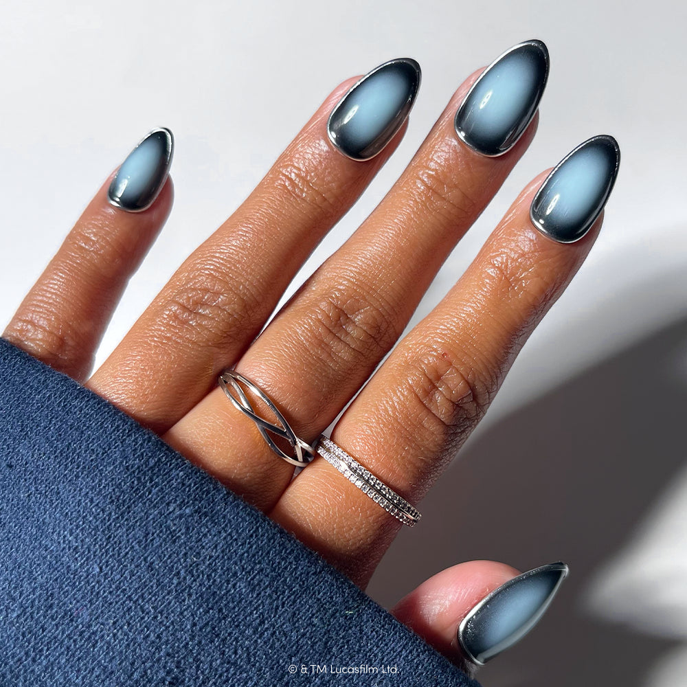 A hand with almond-shaped nails painted in the “Emperor” blue and black gradient from glamnetic is shown against a soft background, adorned with two silver rings. The sleeve of a dark blue garment covers part of the wrist, accentuating the elegance of these Emperor press-on nails.