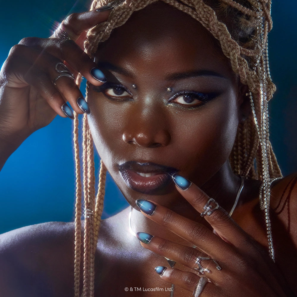 A woman with blonde braids poses with her glamnetic Emperor silver and blue cruelty-free nails near her face, wearing metallic eye makeup, silver rings, and necklaces against a blue background. - Product image