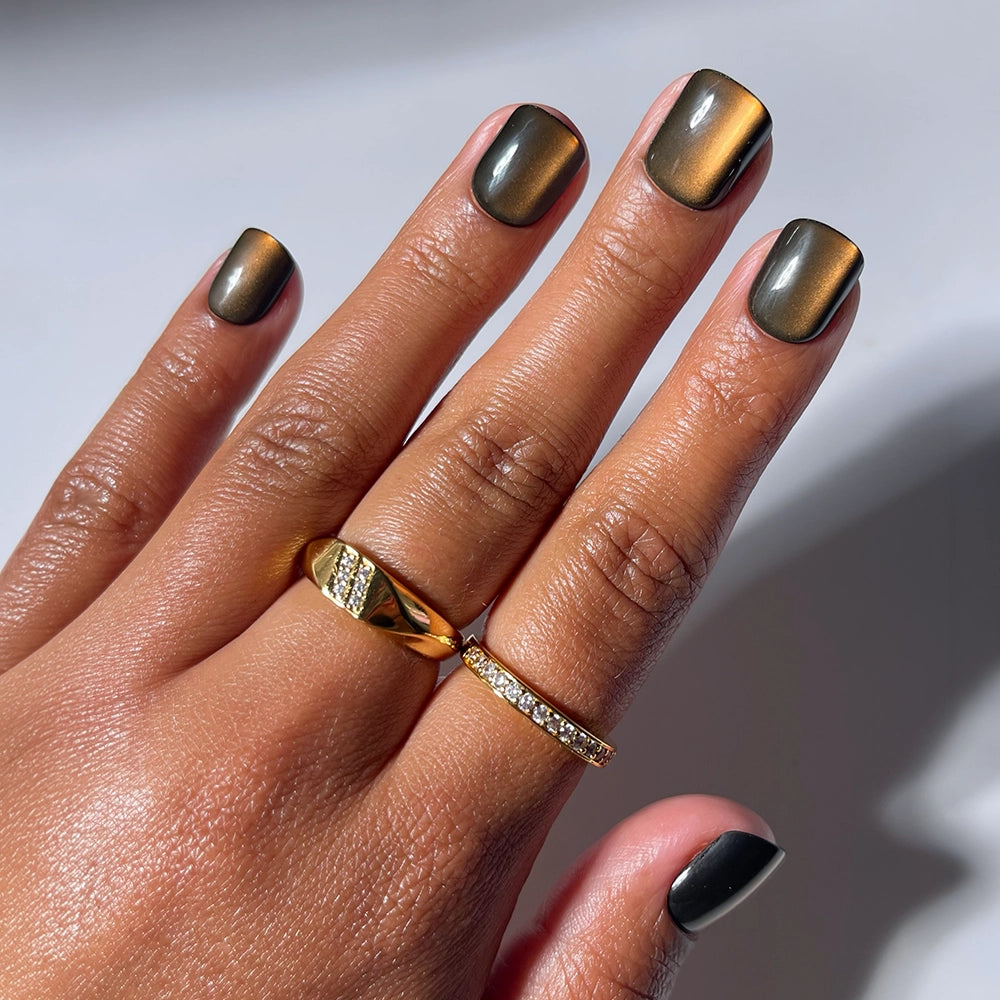 A hand with short, metallic olive-green Enstatite press-on nails by glamnetic rests on a surface. The person wears two gold rings—one solid with a diagonal design and another thin band set with small diamonds. These salon-quality glamnetic Enstatite press-ons add a touch of effortless elegance to the overall look.