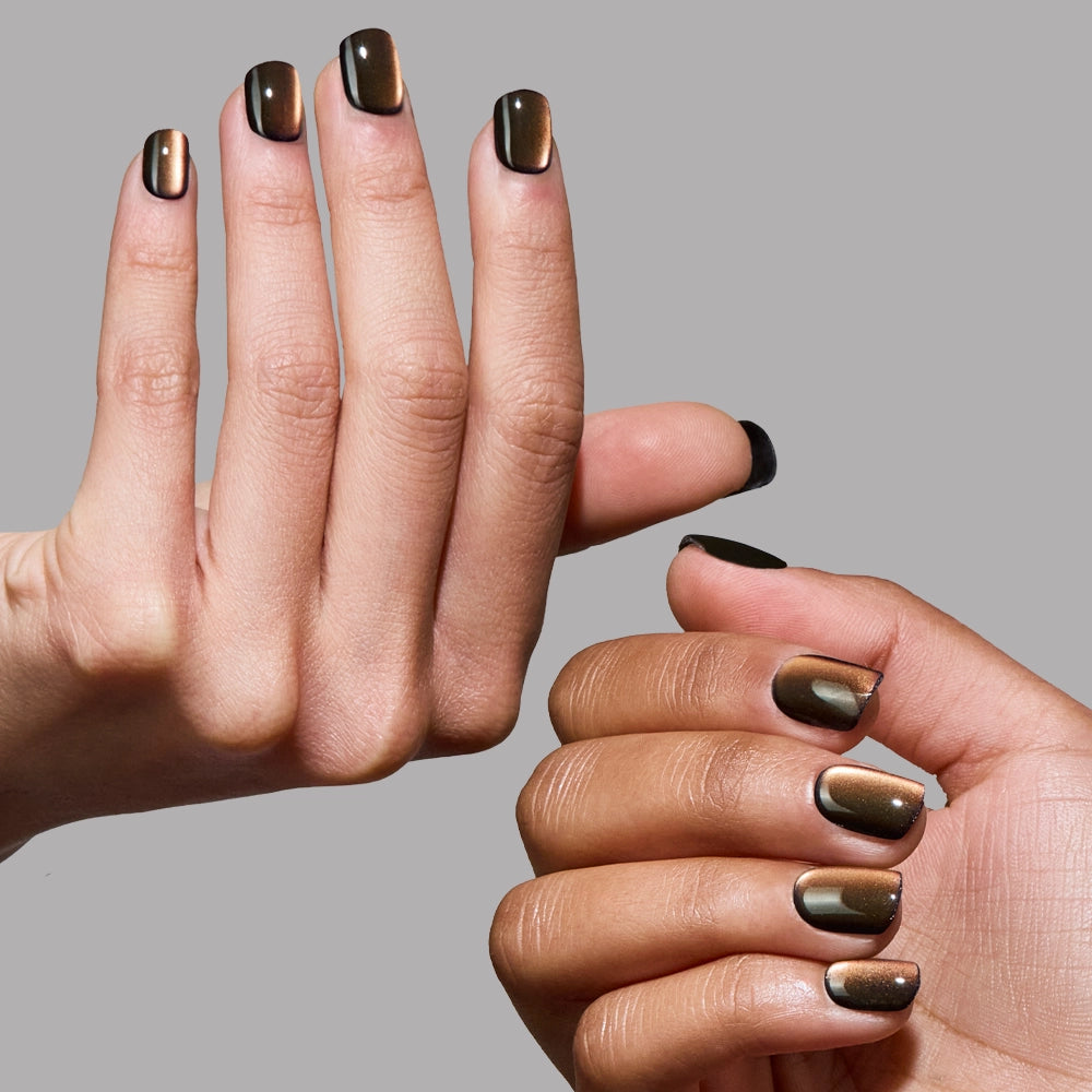 Two hands with glossy, metallic black and bronze gradient nails—just like glamnetic’s Enstatite Press-On Nails—are displayed against a plain gray background.
