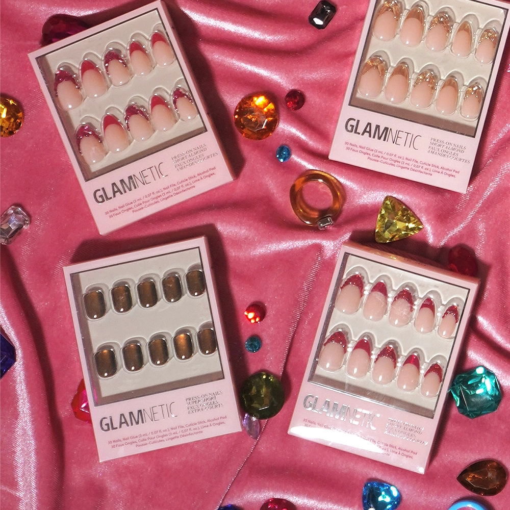 Four boxes of glamnetic press-on nails, featuring the chic Enstatite design, are displayed atop pink satin fabric and encircled by vibrant, faceted gem accents and a statement ring. Each box showcases a unique nail style from glamnetic, delivering salon-quality results for an effortlessly glamorous appearance. - Product image
