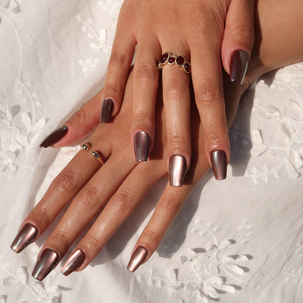 Two hands with medium coffin nails in the metallic rose gold "Espresso Glow" finish by glamnetic rest on white embroidered fabric. The person wears two gold rings—one with red stones and one plain band. - Product image