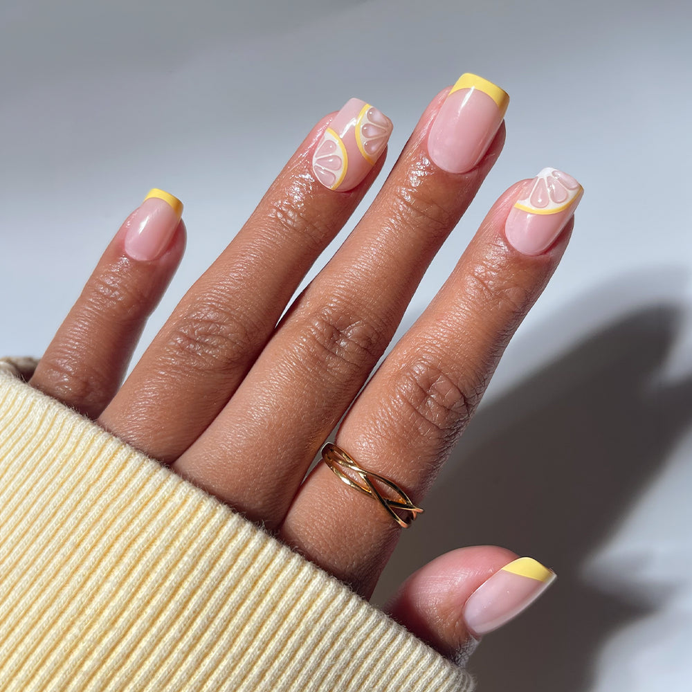 A hand models glamnetic’s Limonata press-ons—short squoval nails in pale pink with yellow French tips and playful lemon art. Worn with a gold ring and yellow ribbed sweater, the look pops against a soft, neutral background.
