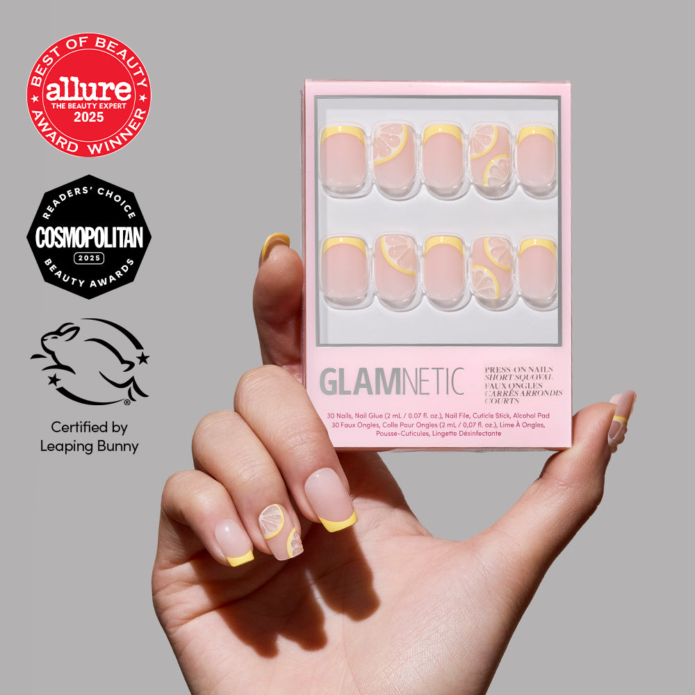 A hand with short squoval yellow French tip press-on nails featuring lemon art holds a pink glamnetic Limonata box, which displays the matching nails and shows beauty award and cruelty-free certification logos.