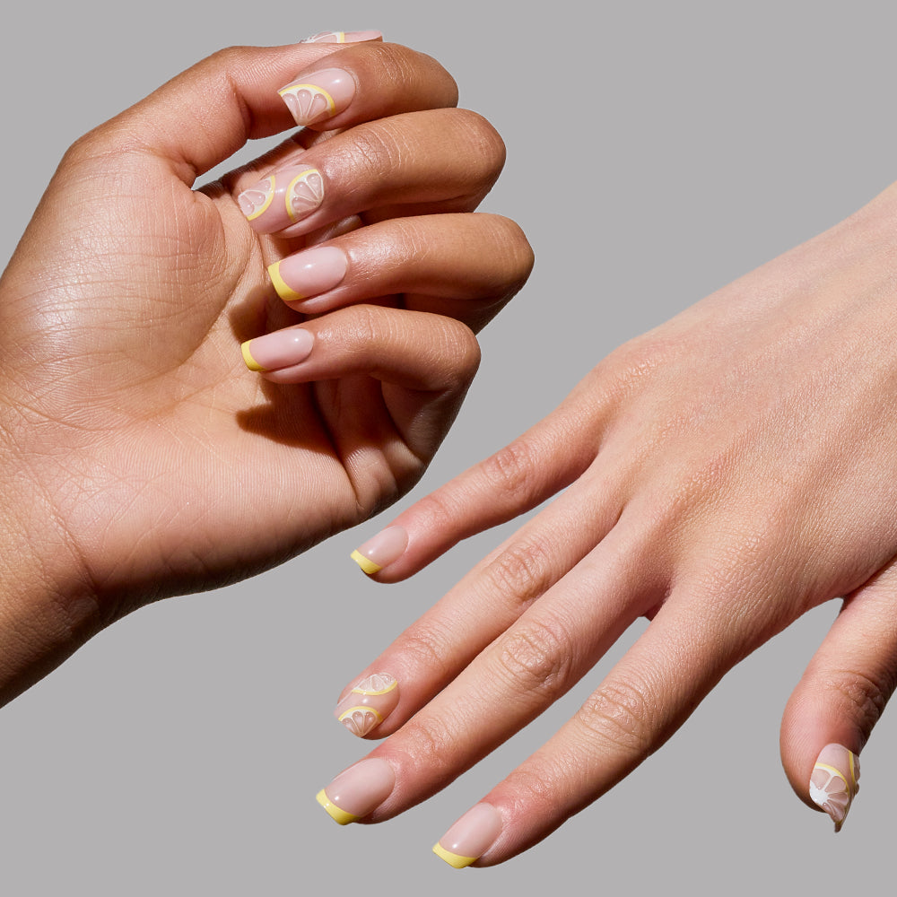 Two hands showcase glamnetic’s Limonata nails: short squoval tips with a natural base, bold yellow French accents, and playful lemon art. The nails are set against a simple light grey background. - Product image