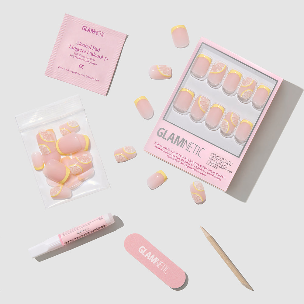 A flat lay displays the glamnetic Limonata press-on nail kit, featuring short squoval nails with pink and yellow lemon art, a nail file, wooden cuticle stick, nail glue, alcohol pad, and extra nails on a light background. - Product image