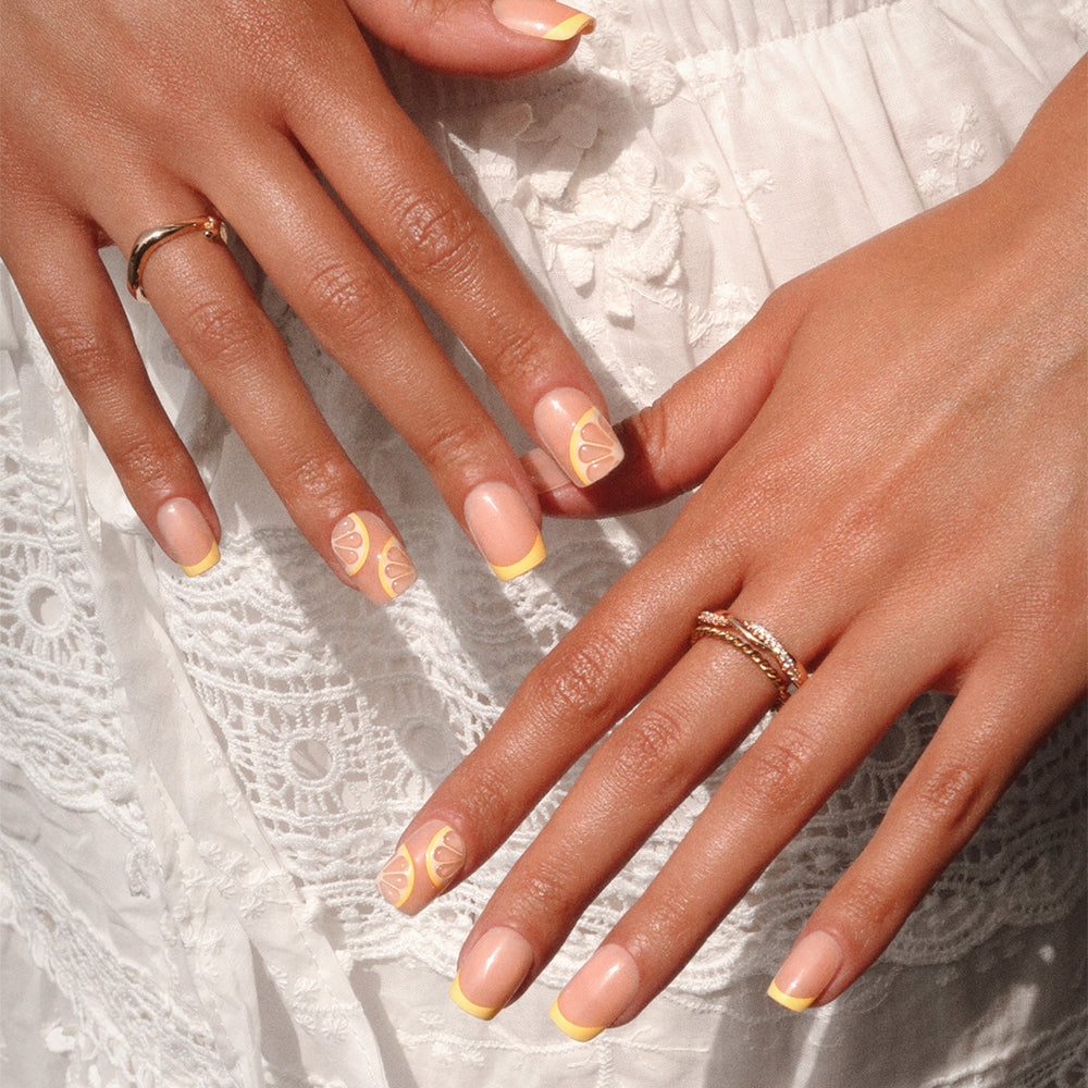 Two hands with rings rest on a white lacy dress, showcasing glamnetic's Limonata nails—short squoval tips with a nude base, yellow French tips, and delicate lemon slice accents for a fresh summer look. - Product image