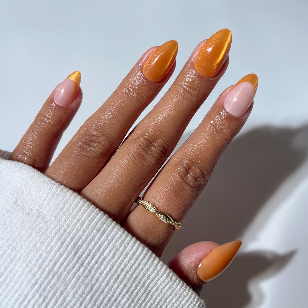 A hand with short almond nails in glamnetic’s Orange Pop press-ons—glossy orange mixed with neutral pink—wears a twisted gold ring and a white sleeve partly covering the wrist.