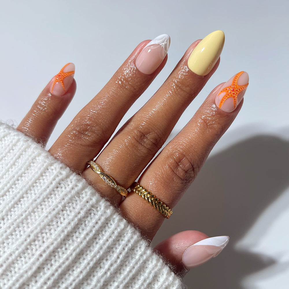 A hand wears glamnetic's Sunset Tide short almond press-ons: orange starfish on index/ring, solid yellow middle, and French tips on thumb/pinky. Gold rings and a white sweater complete the tropical look.