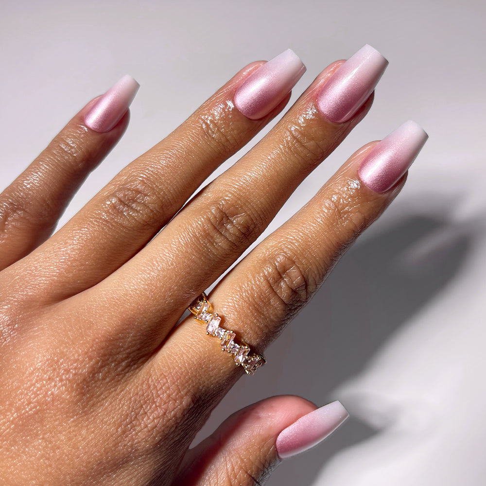 A hand wears glamnetic's EVERYDAY BEAUTY QUEEN press-on nails—long, square-shaped pink and white ombré with a glossy finish—plus a gold ring with clear rectangular gemstones on the ring finger, shown against a light background.