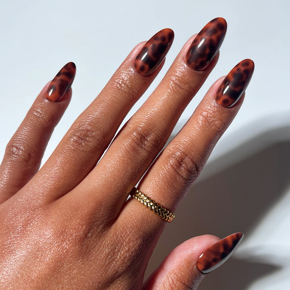 A hand flaunts glamnetic’s Expensive Taste press-on nails—long, almond-shaped with a glossy brown tortoiseshell design—and a gold braided ring on the middle finger, all set against a clean white background. - Product image
