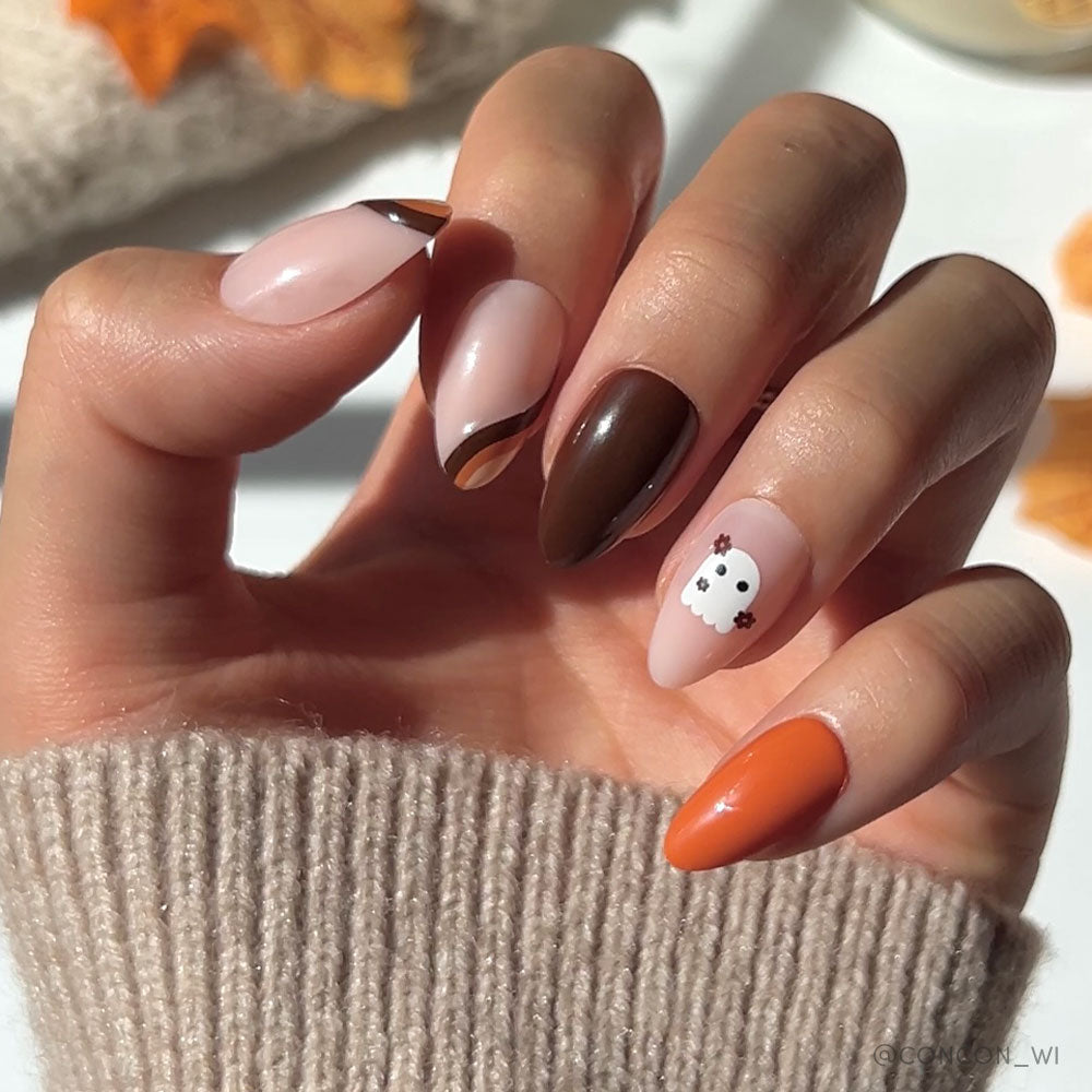 A hand with glamnetic Fall Boo nails—long almonds in brown, beige, and orange, with a playful white ghost accent—perfect for Halloween. The look is paired with a beige knit sweater and autumn leaves in the background.