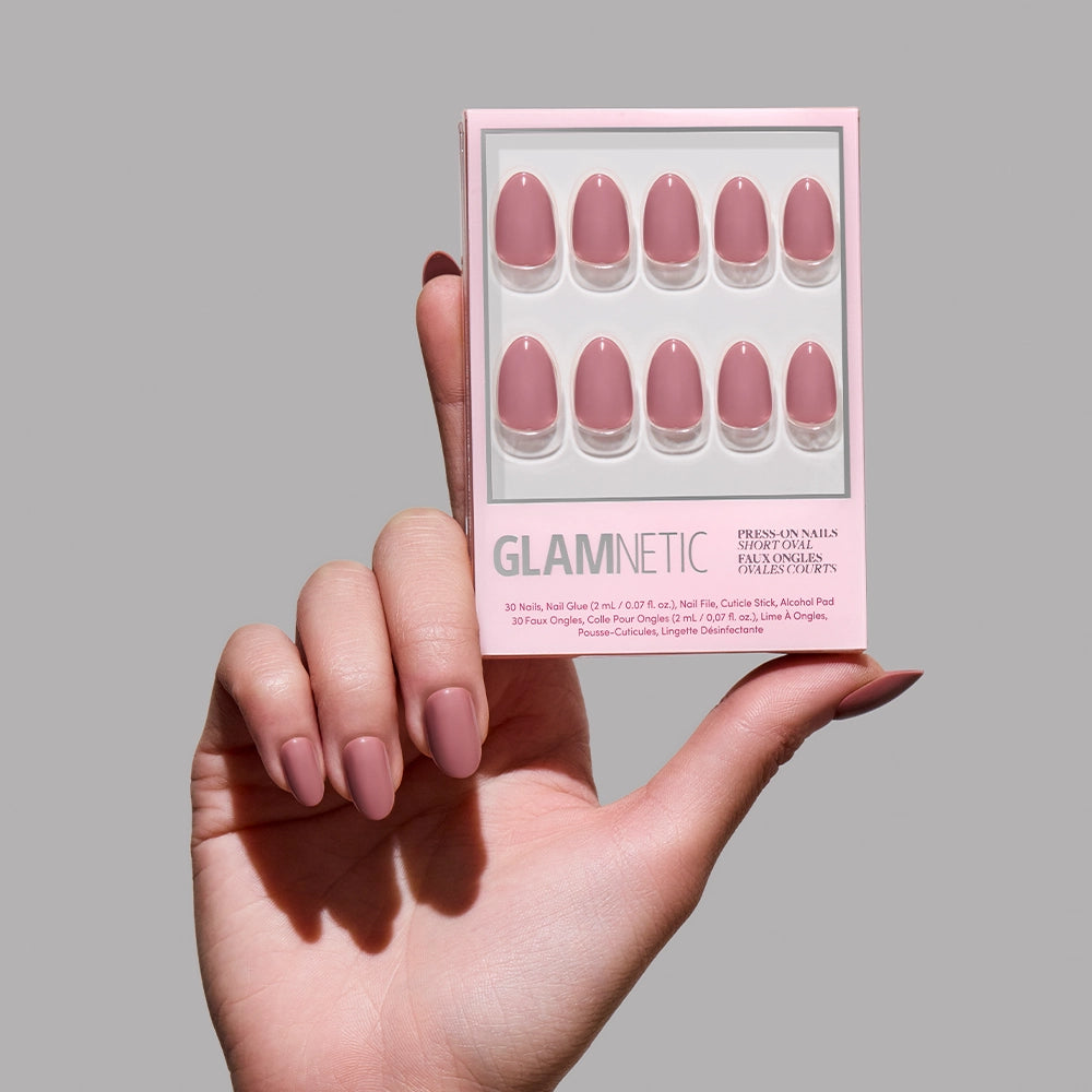 A hand with neatly manicured, mauve nails holds a glamnetic Fall Mauve press-on nail set package in a matching shade against a plain gray background—ideal for highlighting fall nail trends.