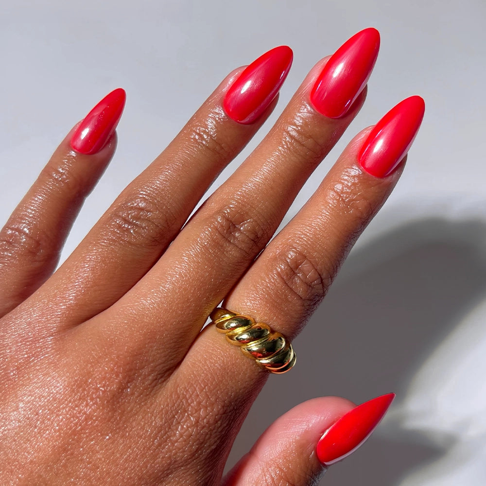 A hand with long, almond-shaped nails painted in glamnetic’s Fearless Red is shown against a white background, the vibrant shade giving a salon-quality look while a chunky gold ring adorns the middle finger.