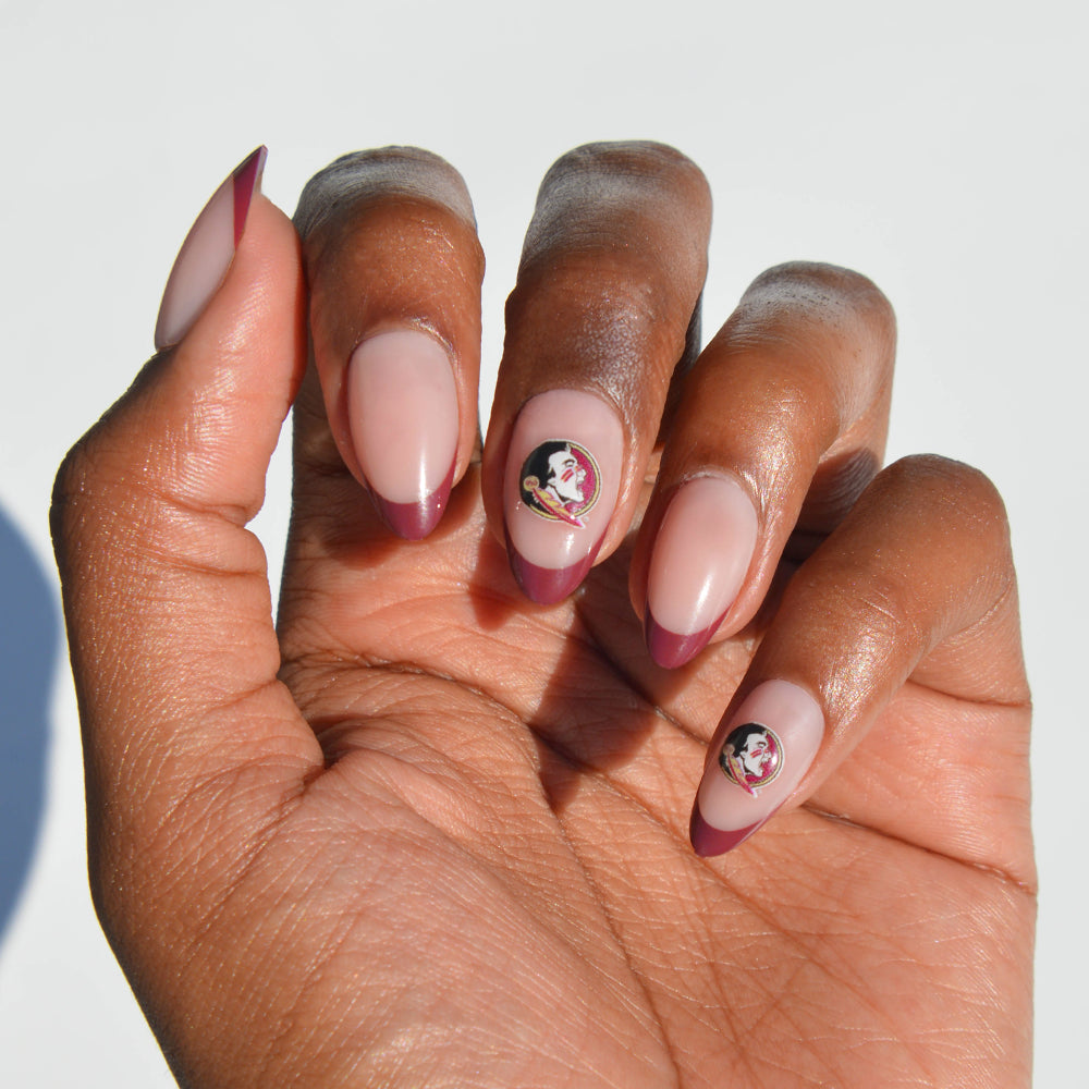 A hand with almond-shaped nails showcases glamnetic’s Collegiate Press-On Nails in a nude base with maroon tips; two nails are detailed with a small face and feather logo, resembling a sports team emblem. - Product image