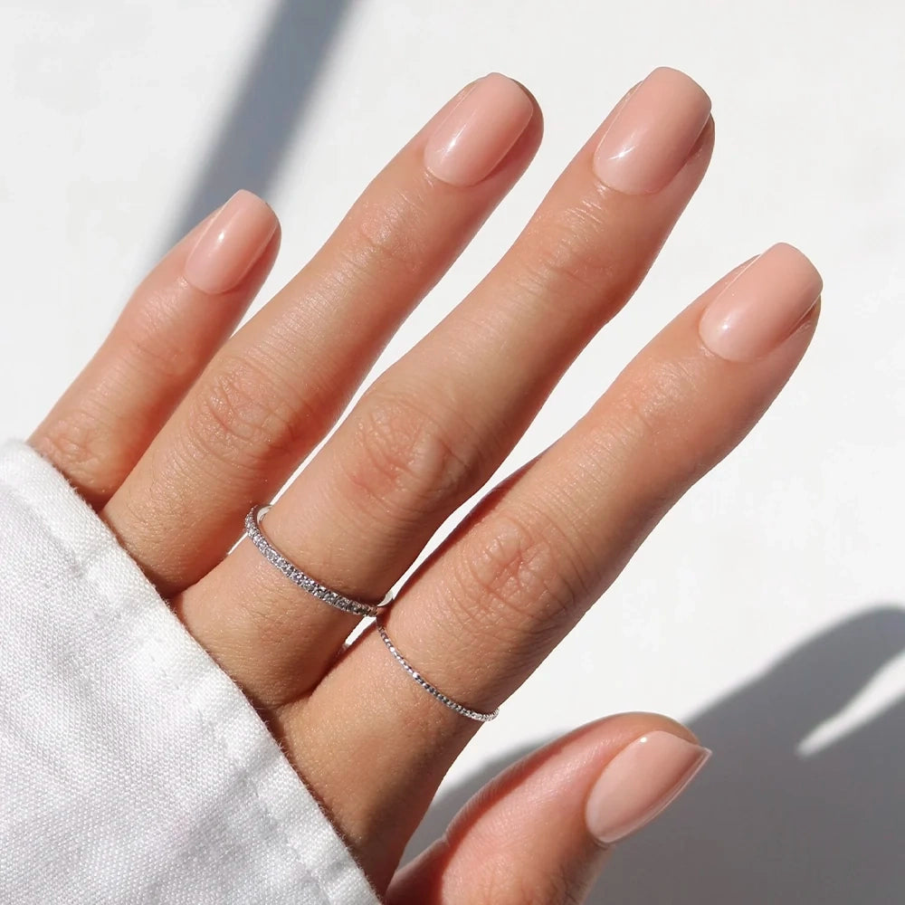 A hand with neatly manicured, short nude nails—created using glamnetic’s FOR THE “IT” GIRLS—rests gracefully. The person is wearing two thin silver rings on the middle and ring fingers, and the sleeve of a white shirt is partially visible, highlighting the salon-quality finish of these chic press-on nails.