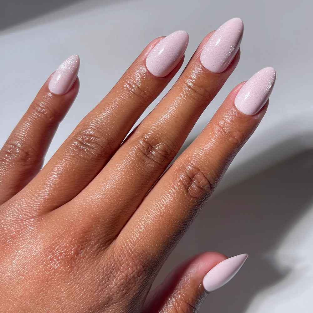 A hand with medium brown skin wears glamnetic's Forever Pink press-on nails, featuring long, almond shapes in a glossy pale pink shade. The well-manicured nails have a smooth finish and a subtle shimmer under light.