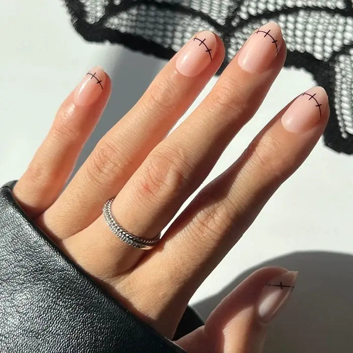 A hand displays short, nude-colored nails from the glamnetic Frankenbabe set, featuring minimalist black stitch-inspired lines near the tips for a chic Halloween twist. The look is complemented by a silver ring and a black leather sleeve.
