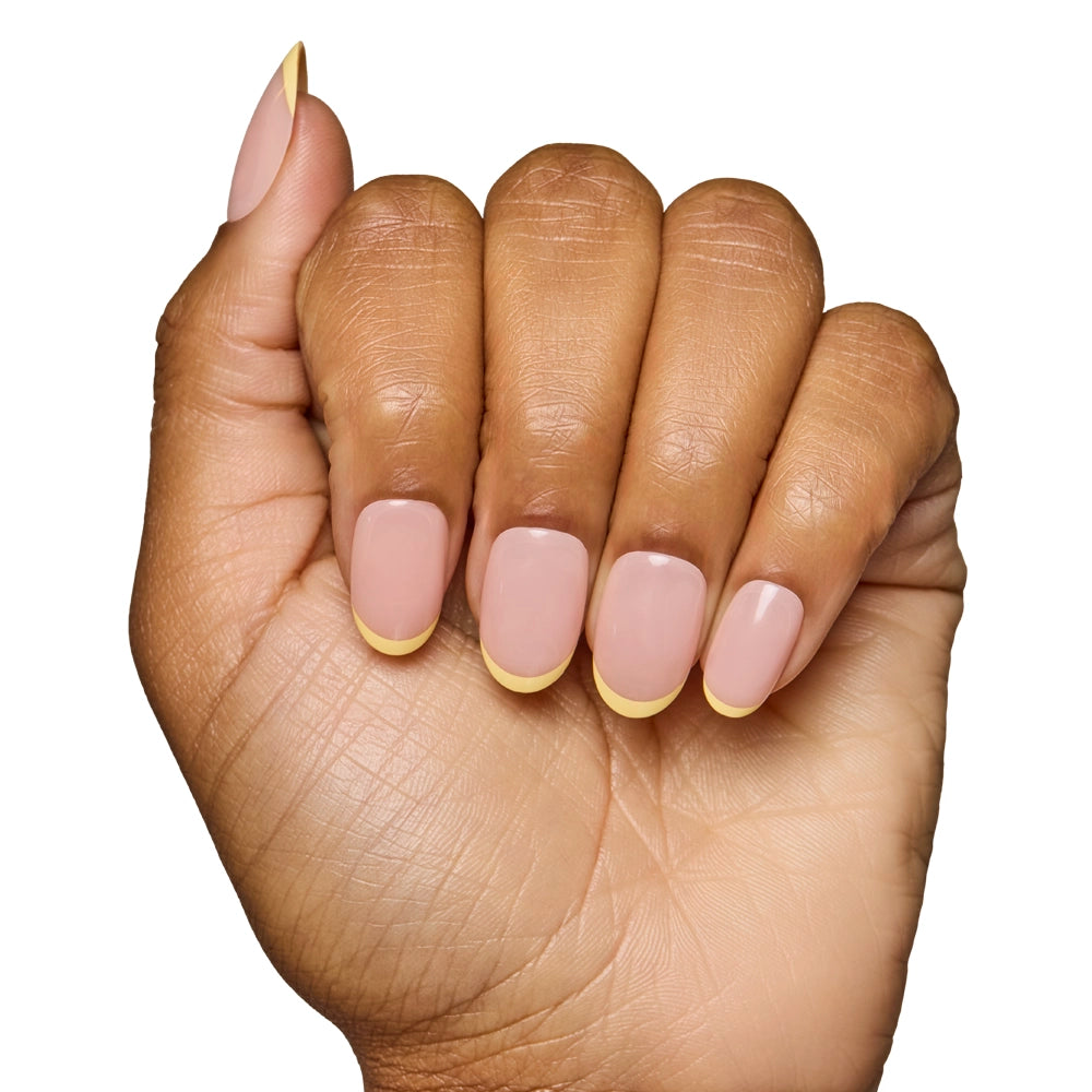 A hand with medium-brown skin displays the glamnetic French Butter Press-On Nail Set—nude base with pastel yellow French tips—against a white background, highlighting its salon-quality durability and stylish look. - Product image