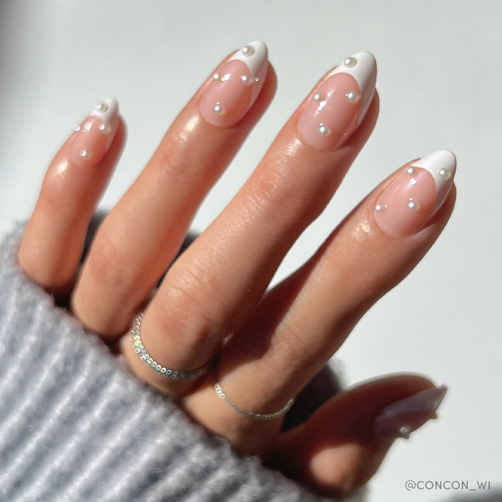 A hand with glamnetic’s French Pearl features short almond nails with a sheer pink base, white French tips, and pearl-like accents—ideal for bridal events. The person accessorizes with silver rings and a ribbed light gray sweater. - Product image