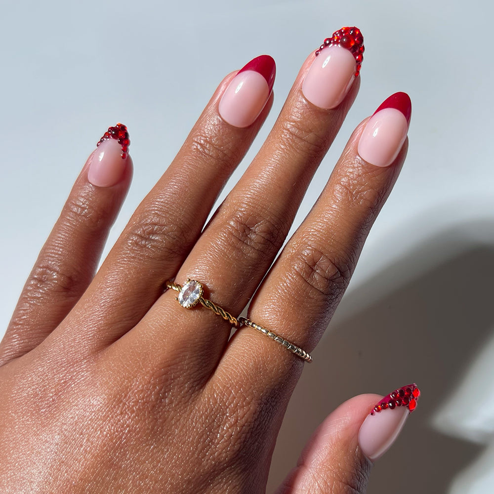 A hand showcases glamnetic’s Garnet nails, featuring almond-shaped tips with a pink base, vibrant red French tips, and sparkling red rhinestone accents. The look is complemented by two gold rings, one set with a clear oval stone.