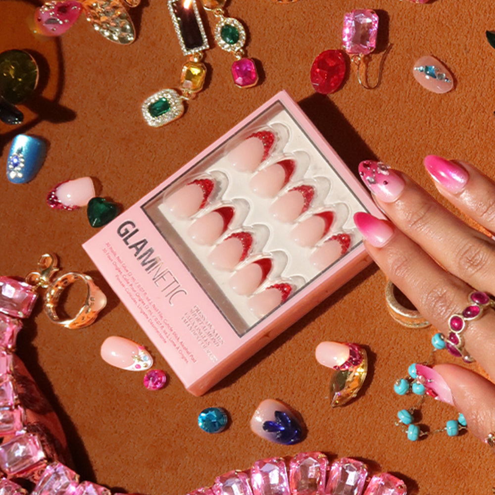 A hand with pink and glittery nails rests near a box of glamnetic Garnet press-on nails, surrounded by colorful rings, gems, jewel-encrusted nails, and decorated false nails on a brown surface. - Product image