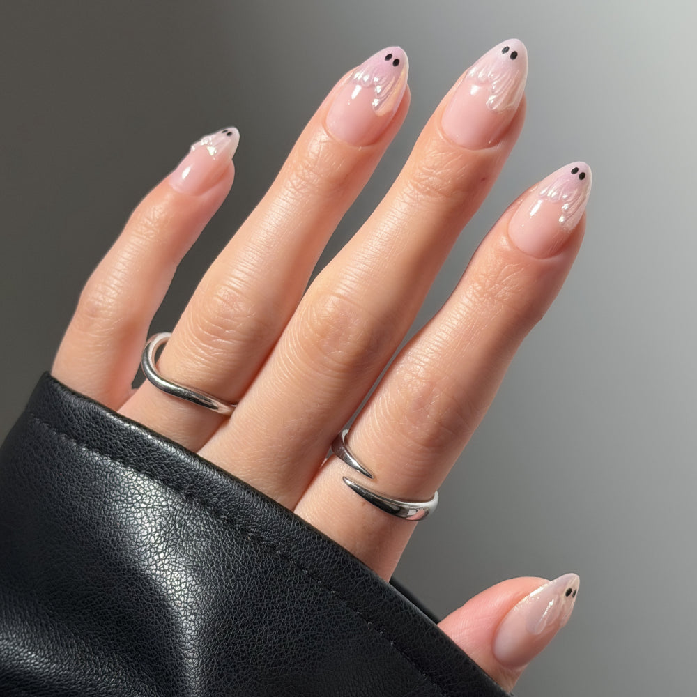 A hand with short almond nails showcases glamnetic’s Ghost Besties press-ons, featuring abstract white and black ghost nail art. The person wears silver rings, with a black leather sleeve partially covering the hand.