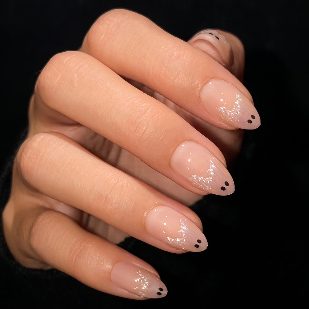 A hand with short almond nails painted in glamnetic’s Ghost Besties, a sheer nude polish, featuring simple black dot designs near the tips. The lighting highlights the shine of the manicure against a black background. - Product image