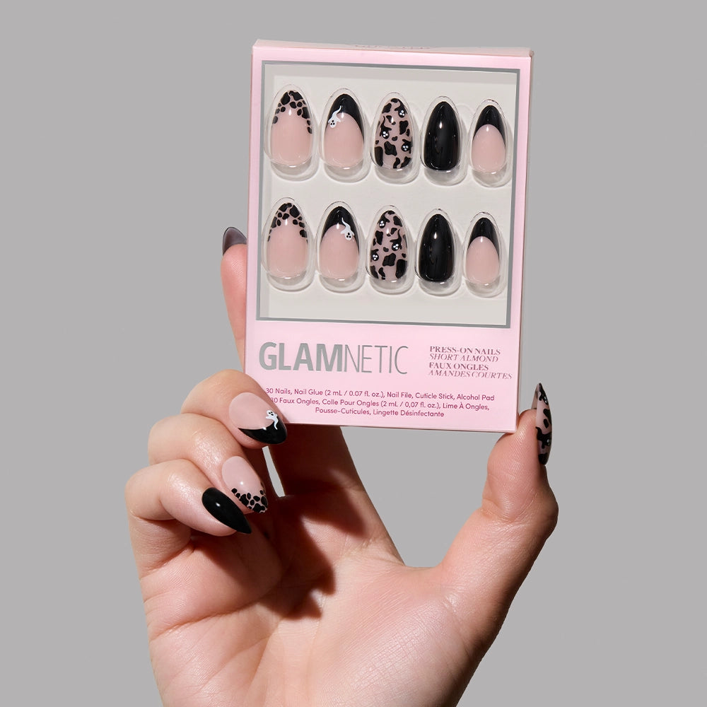 A hand with long, black and leopard print press-on nails holds a package of glamnetic Ghosted press-on nails featuring similar designs against a plain gray background.