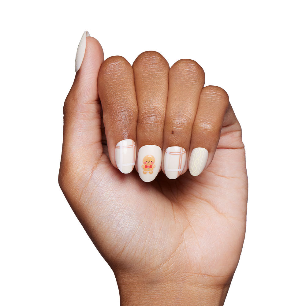 A hand with glamnetics Gingerbread Knit press-on nails features beige polish, festive holiday designs like plaid patterns, and a gingerbread man illustration on the middle nail—making it the perfect accessory against a white background. - Product image