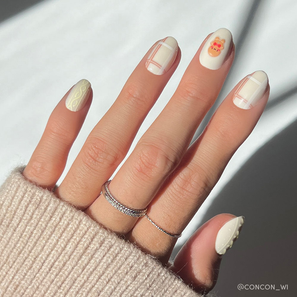 A hand with cream geometric nail art, a cute cartoon bear, and textured swirls—styled with thin rings and a beige sweater—makes the perfect festive look when paired with glamnetics Gingerbread Knit nail set.
