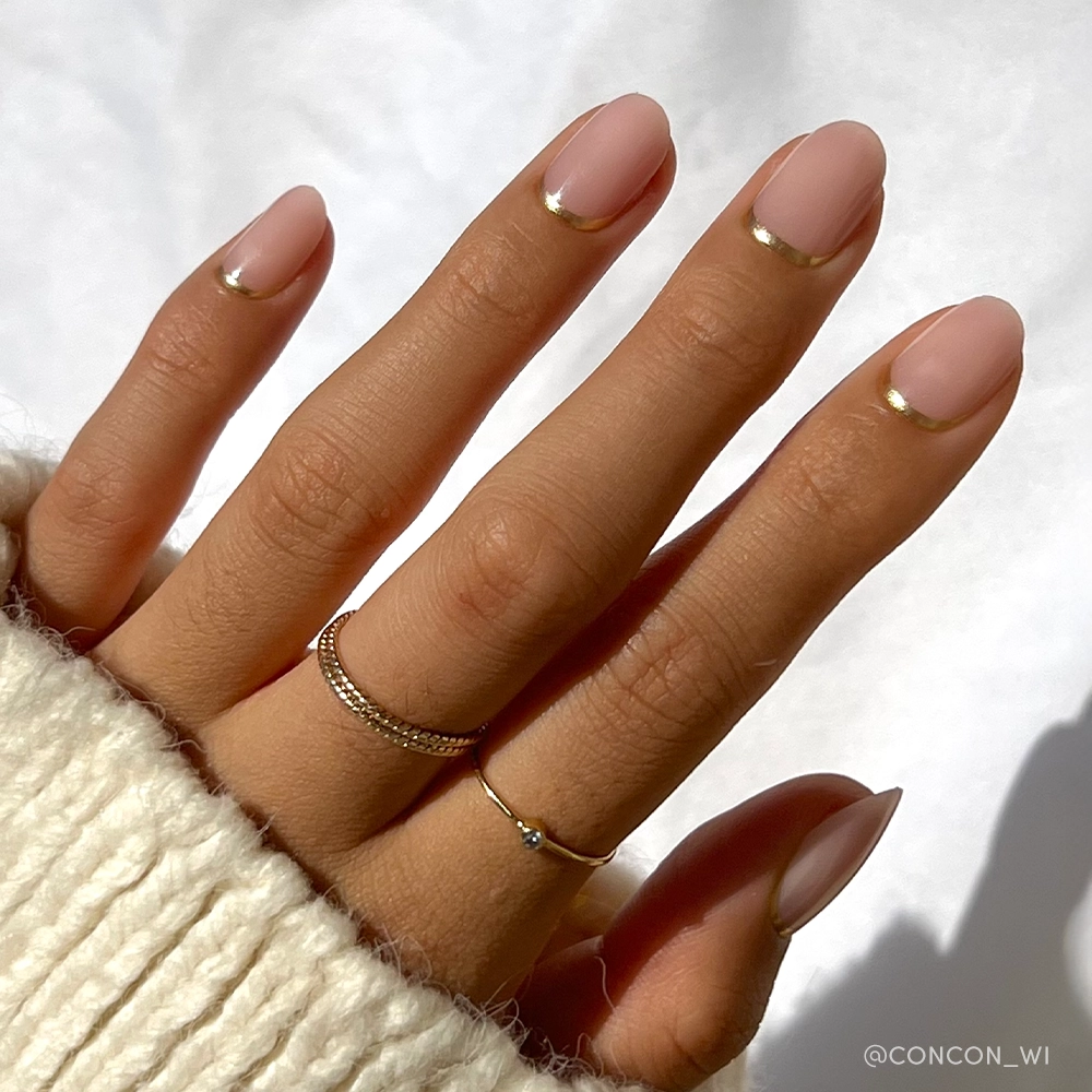 A hand with short round nails in glamnetic&#39;s Goal Digger, featuring a sheer pink base and thin metallic gold tips. The person wears a cream sweater and two delicate rings—one on the ring finger, one on the pinky.