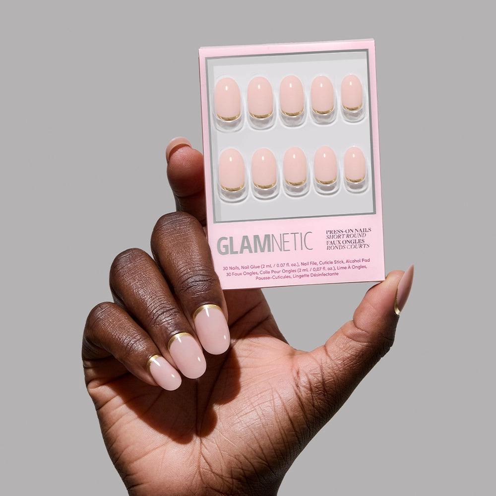 A hand with short, round, light pink nails with gold foil (Glamnetic Goal Digger) holds the matching press-on nail box by glamnetic against a plain gray background.