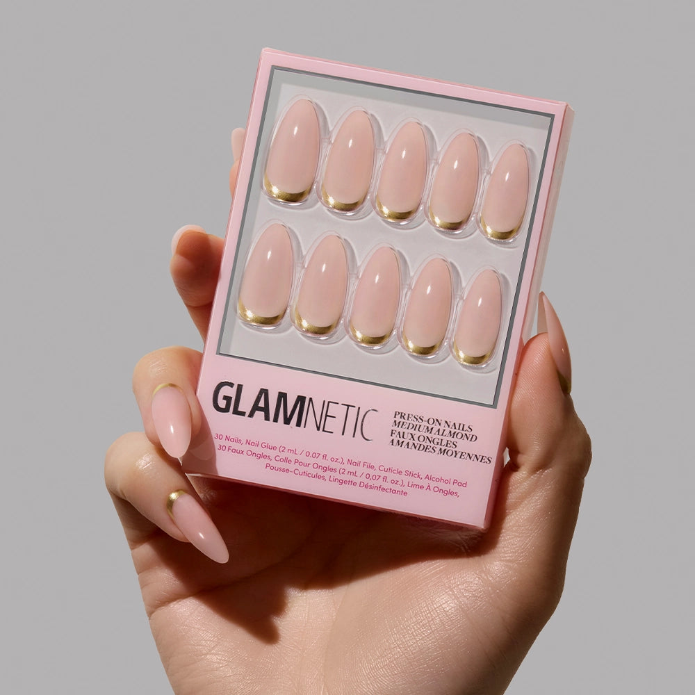 A hand with medium almond nude pink nails holds a box of glamnetic Goal Getter press-on nails, featuring elegant gold French tips, against a plain gray background.