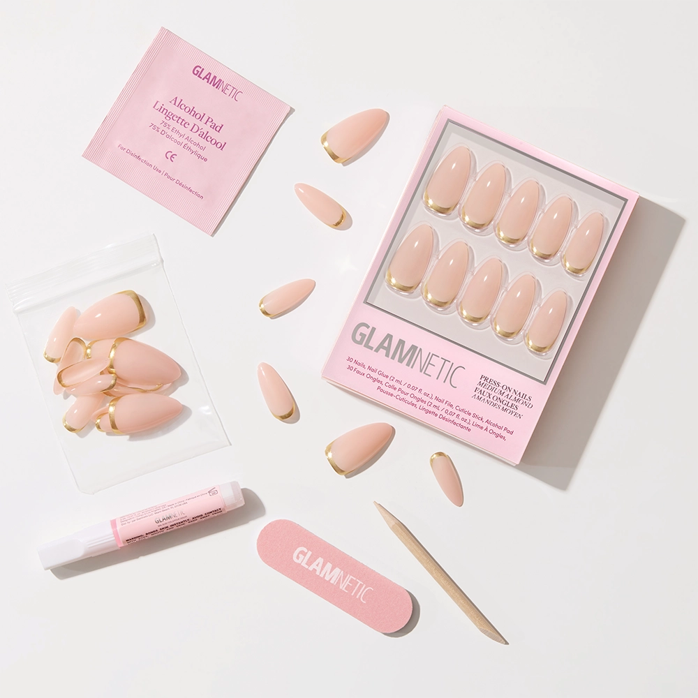 The glamnetic Goal Getter press-on nail kit includes nude almond-shaped nails with gold French tips, adhesive, a pink nail file, cuticle stick, alcohol pad, and spare nails—all arranged neatly on a white background. - Product image