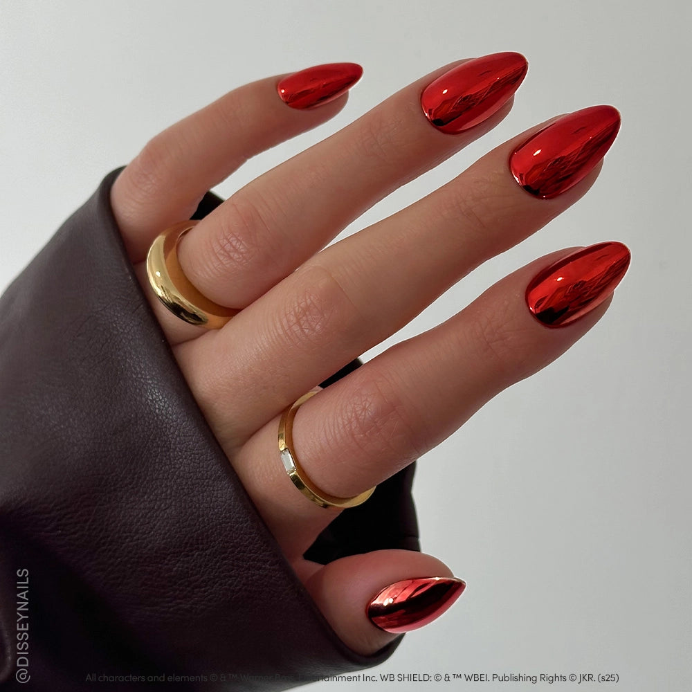 A hand with long, almond-shaped Godric Gryffindor™ red chrome nails by glamnetic glows in metallic polish. Two gold rings adorn the fingers, and a dark brown sleeve partially covers the hand. - Product image