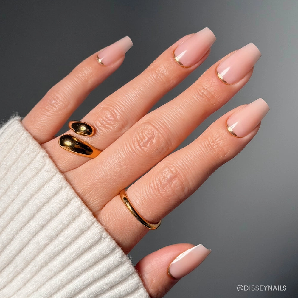 A hand with square-shaped nude pink Gold Goals Press-On Nails by glamnetic, featuring thin gold crescent accents near the cuticles, is accessorized with three gold rings and a cream ribbed sleeve for a chic salon-quality look.