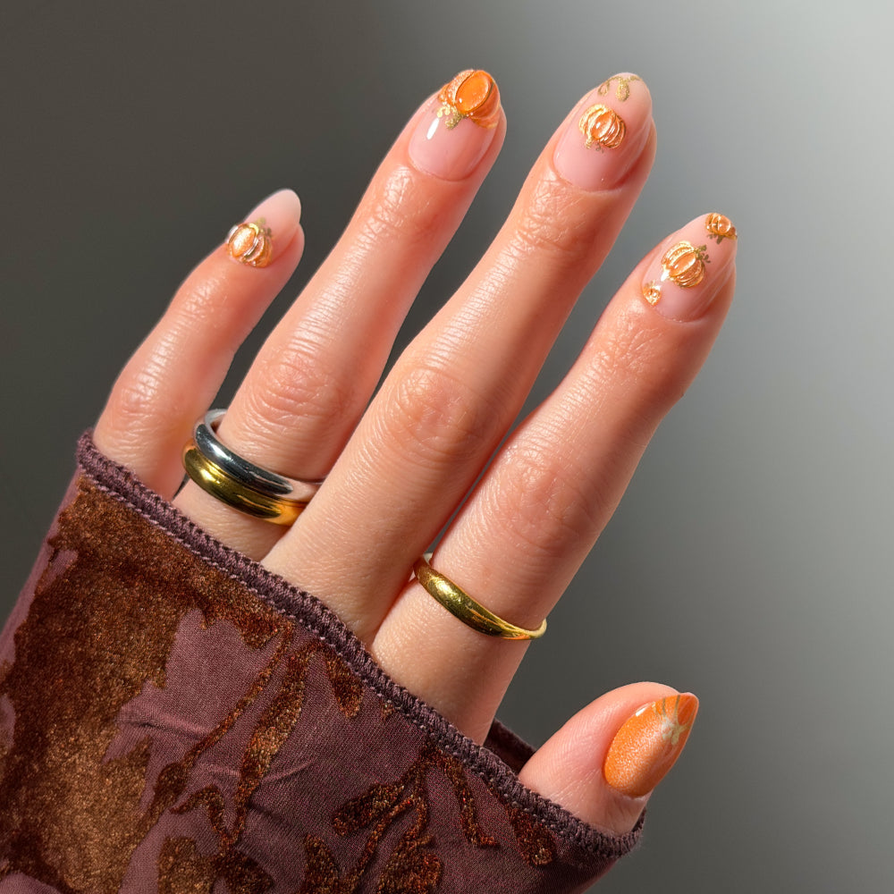 A hand adorned with gold and silver rings and dressed in a brown velvet sleeve showcases glamnetic’s Golden Gourds nails—short oval tips manicured in glossy orange polish with gold rose designs, featuring elegant pumpkin-inspired nail art and a shimmery orange accent on the thumb.