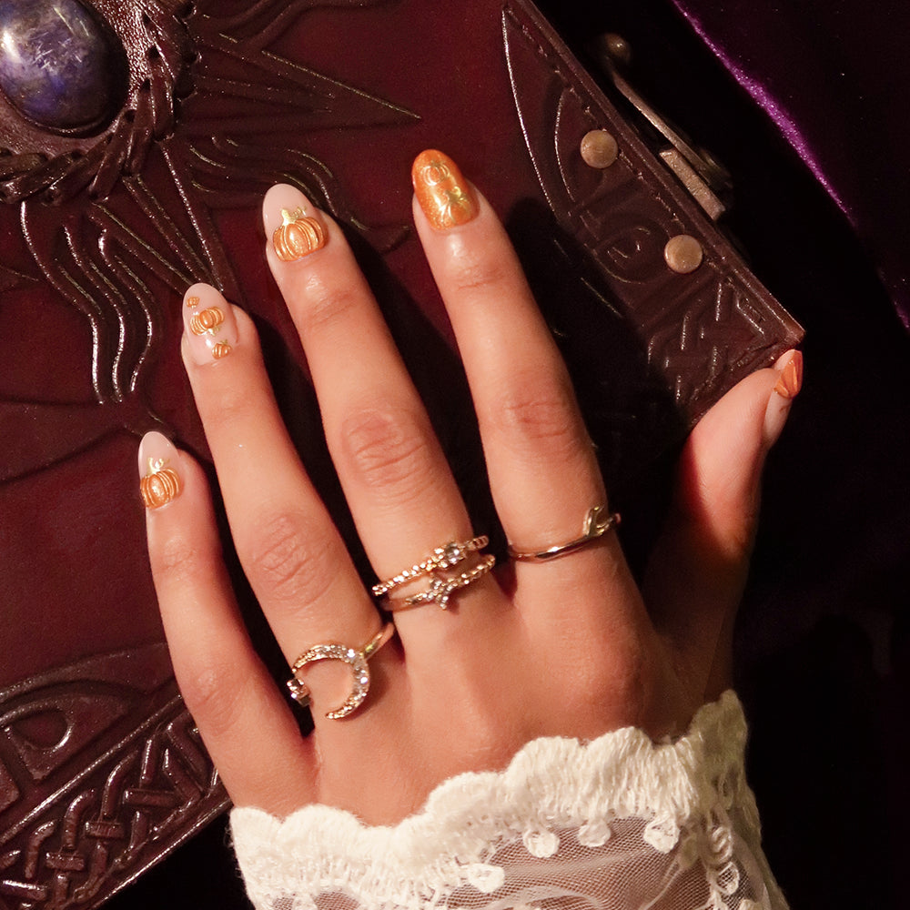 A close-up of a hand adorned with glamnetic’s Golden Gourds gold moon and star rings, showcasing manicured short oval nails detailed with delicate pumpkin nail art. The hand rests on a dark, ornate book featuring a blue stone, set against a deep purple background, with a white lace sleeve in view. - Product image