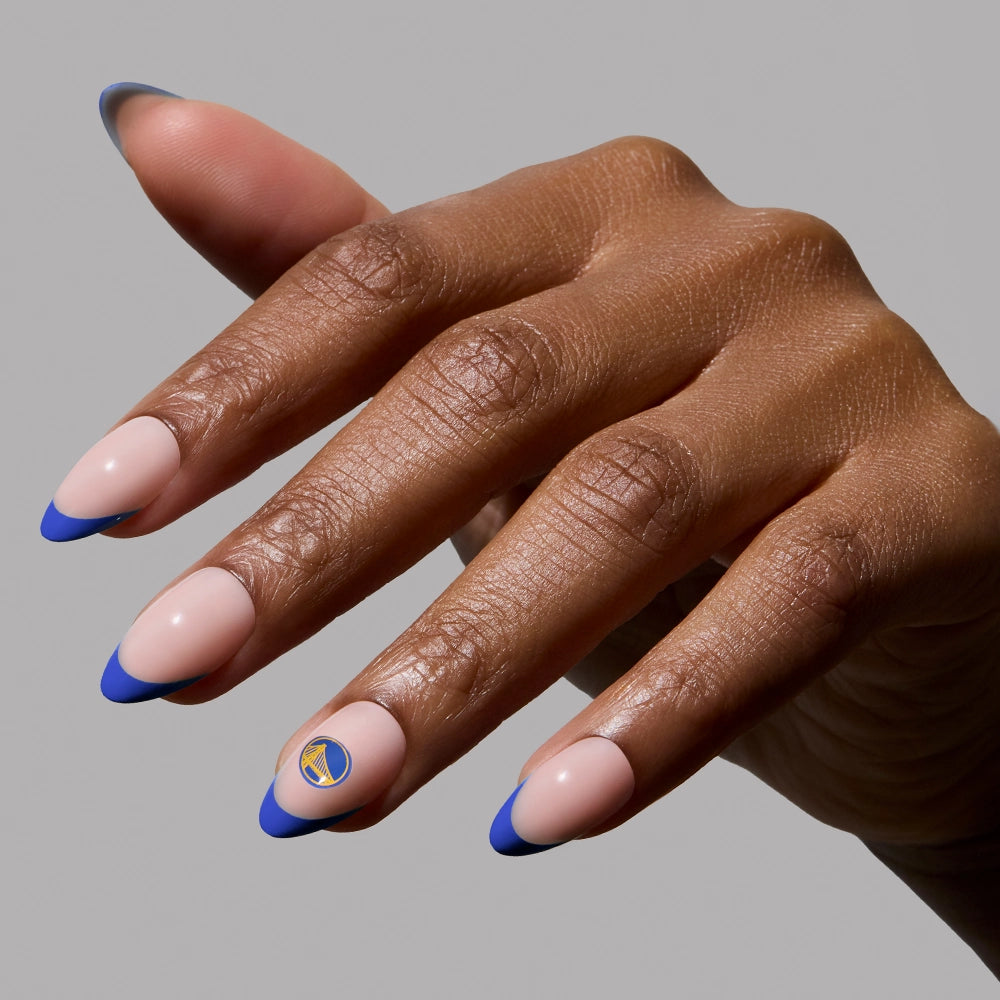 A hand with French tip almond nails—nude base and blue tips—features glamnetic’s Golden State Warriors Press-On Nail Set, including a blue and gold bridge emblem. The background is plain light gray.