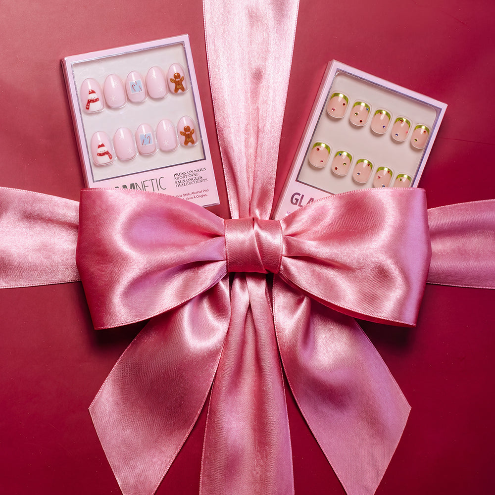 Two boxes of Goodie Gumdrops vegan press-on nails featuring gingerbread and holiday-themed designs sitting on a red surface behind a large pink satin bow. - Product image