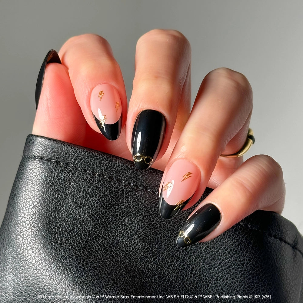 A hand with glamnetic Harry Potter™ almond press-on nails shows a black and nude mani with gold lightning bolt and star details, holding a textured black clutch—perfect for fans seeking magical, themed nails. - Product image