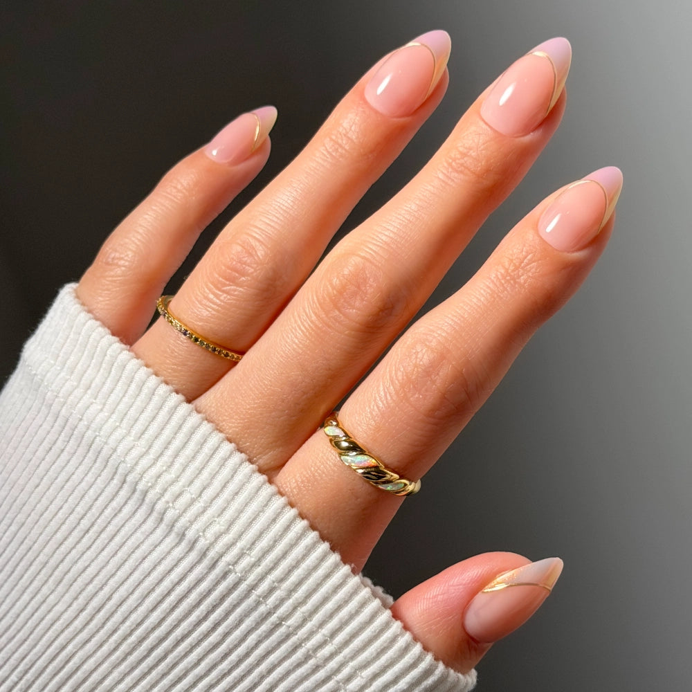 A hand with long, almond-shaped nails in the light pink “Heavenly Touch” design by glamnetic, featuring subtle chrome French tips and two gold rings, is partially covered by a white ribbed sleeve. - Product image