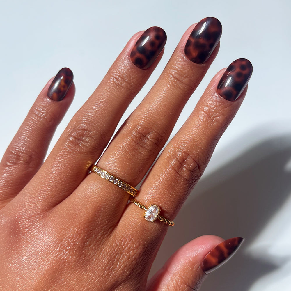 A hand with almond-shaped, brown tortoiseshell "Hidden Fortune" manicure nails by glamnetic, adorned with two gold rings—one set with small diamonds and the other featuring an oval gem—shown against a white background.
