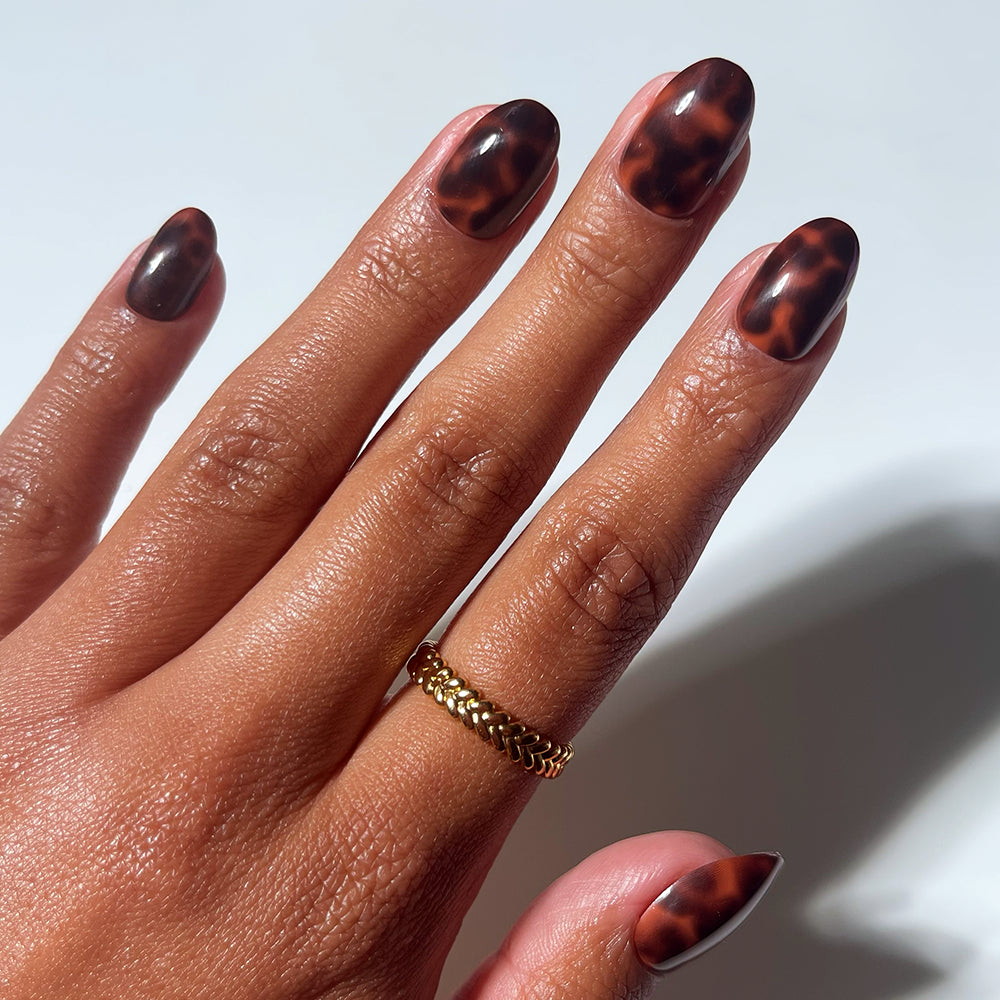 A hand with almond-shaped High Society nails by glamnetic in a brown tortoiseshell design, wearing a gold chain ring on the middle finger, displayed against a white background.