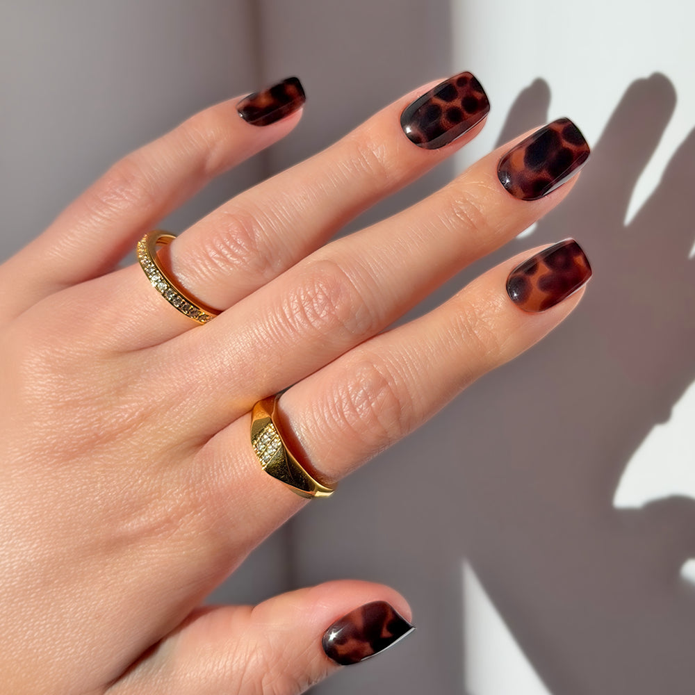 A hand displays medium-length, salon-quality tortoiseshell nails in glamnetic’s High Stakes design, accented by two gold gemstone rings. Natural sunlight casts soft shadows on a white surface.